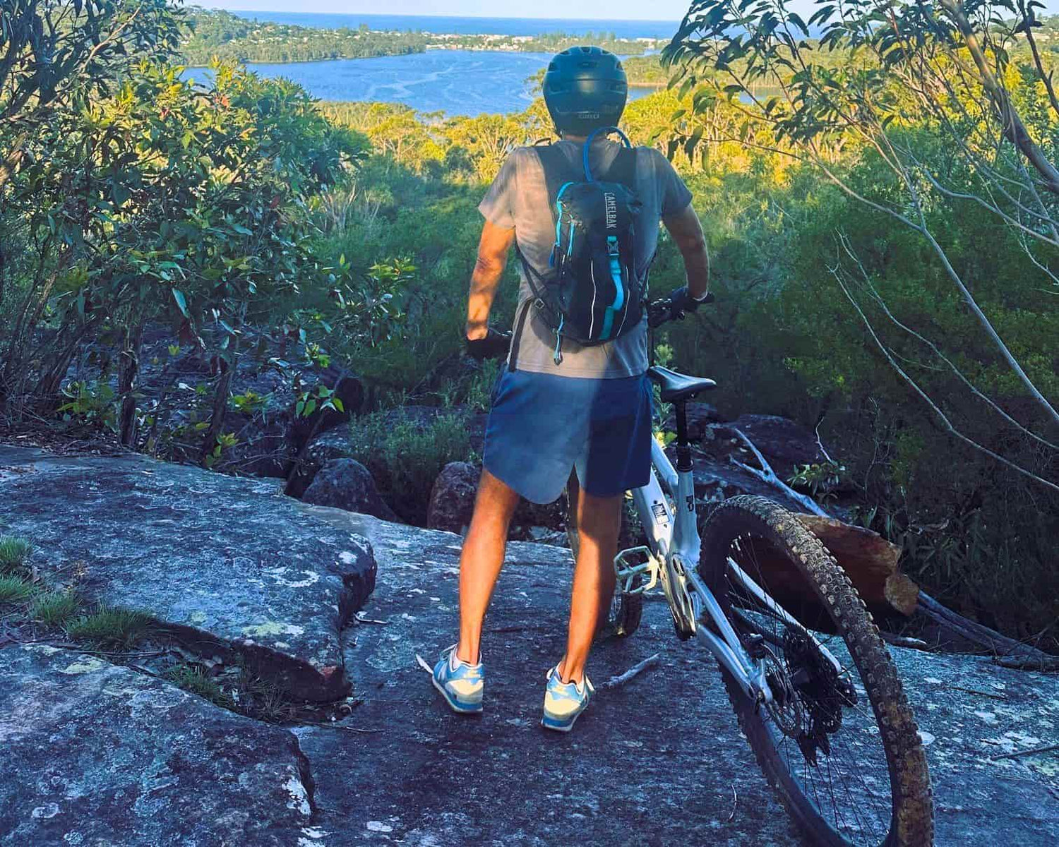 Person with a bicycle on a rocky trail with a scenic view of water and trees.