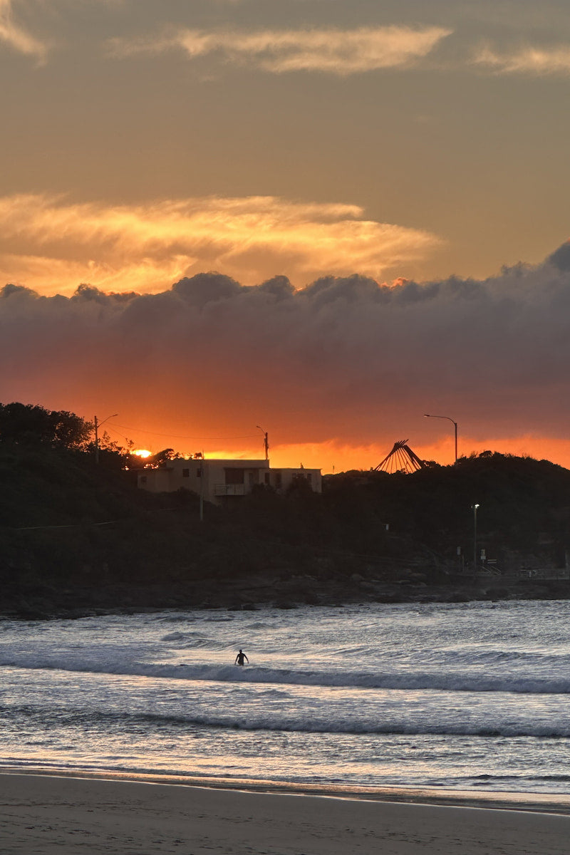Sunrise over Freshwater on Sydney's Northern Beaches with a person in the water and silhouettes of trees and buildings.